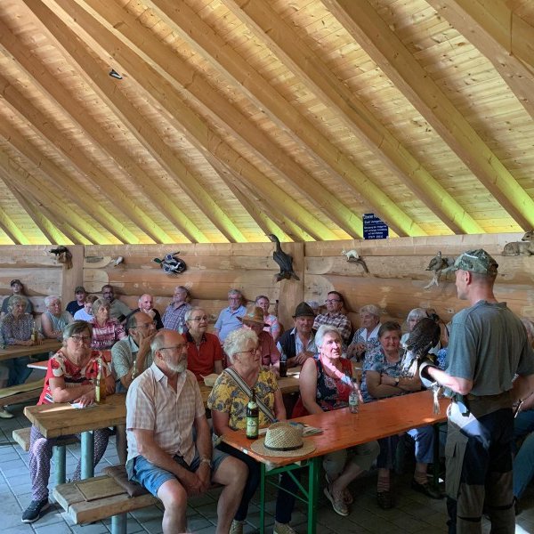 Mann hält einen Vogel und spricht zu einer sitzenden Gruppe älterer Menschen in einem Holzgebäude mit Holzbänken und -tischen, dekoriert mit Tierpräparaten. Text: „Please ask first before taking any photos.“
