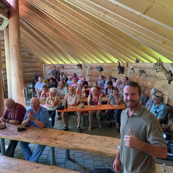 Ein Mann zeigt Daumen hoch. Er steht in einem holzgetäfelten Raum mit vielen älteren Menschen, die an Tischen sitzen und applaudieren. Die Atmosphäre wirkt freudig und gemeinschaftlich.