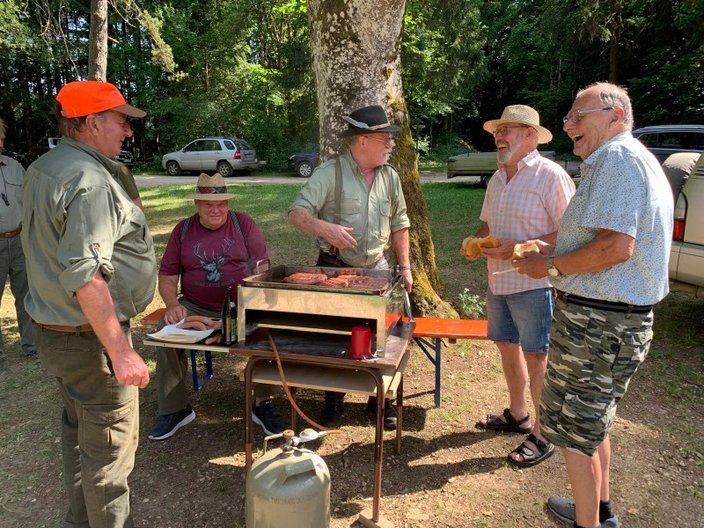 Männer stehen um einen Grill, auf dem Fleisch gegrillt wird; im Hintergrund sind Bäume und geparkte Autos zu sehen. Ein Mann hält Brötchen, während ein anderer kocht.